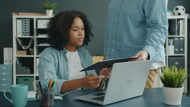 A colleague handing a folder of documents to a coworker at a desk with a laptop, representing audit evidence.
